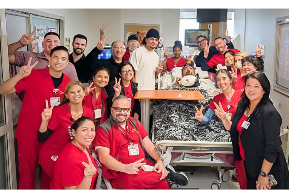 patient in bed with man next to him in cap and nurses in red surrounding the bed
